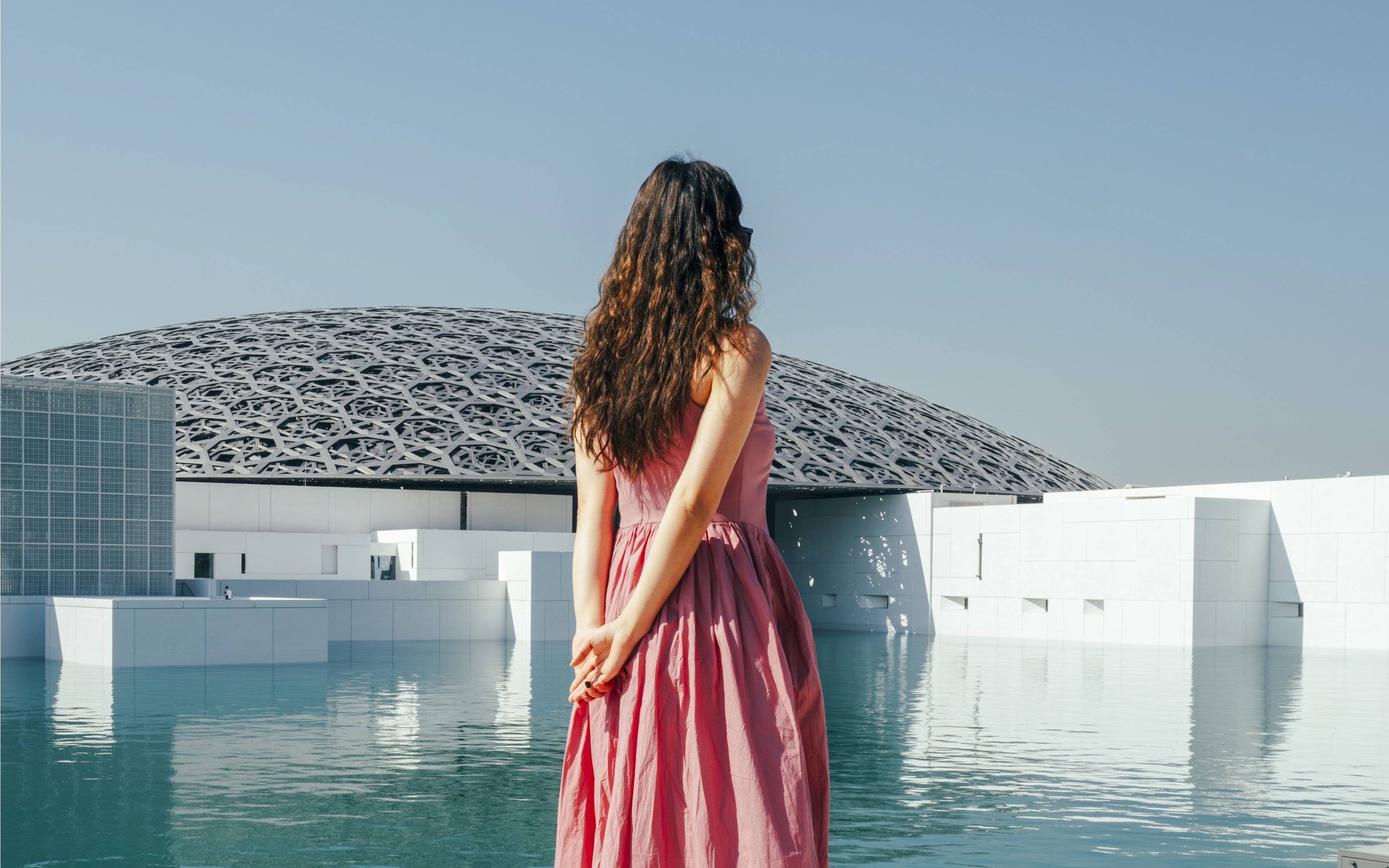 Woman standing by the water at Louvre Abu Dhabi with dome in background.