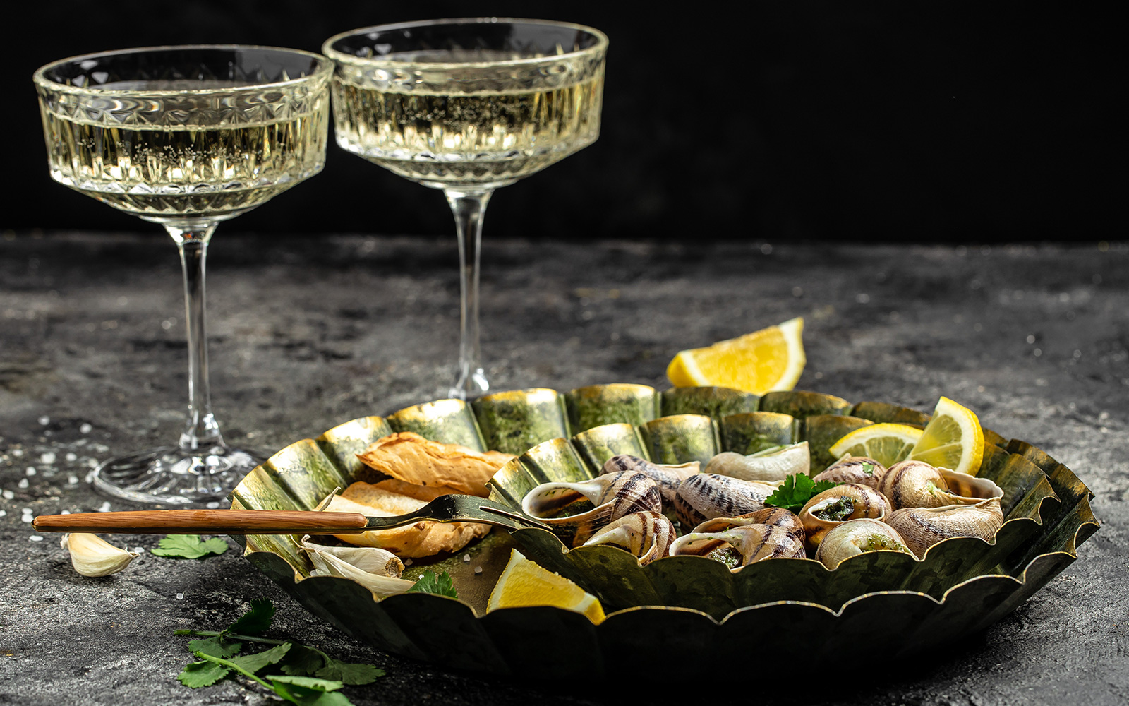 Escargot with lemon wedges and bread on a plate, accompanied by two glasses of white wine.