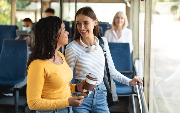 Women smiling and chatting on a bus ride, holding coffee and phone.