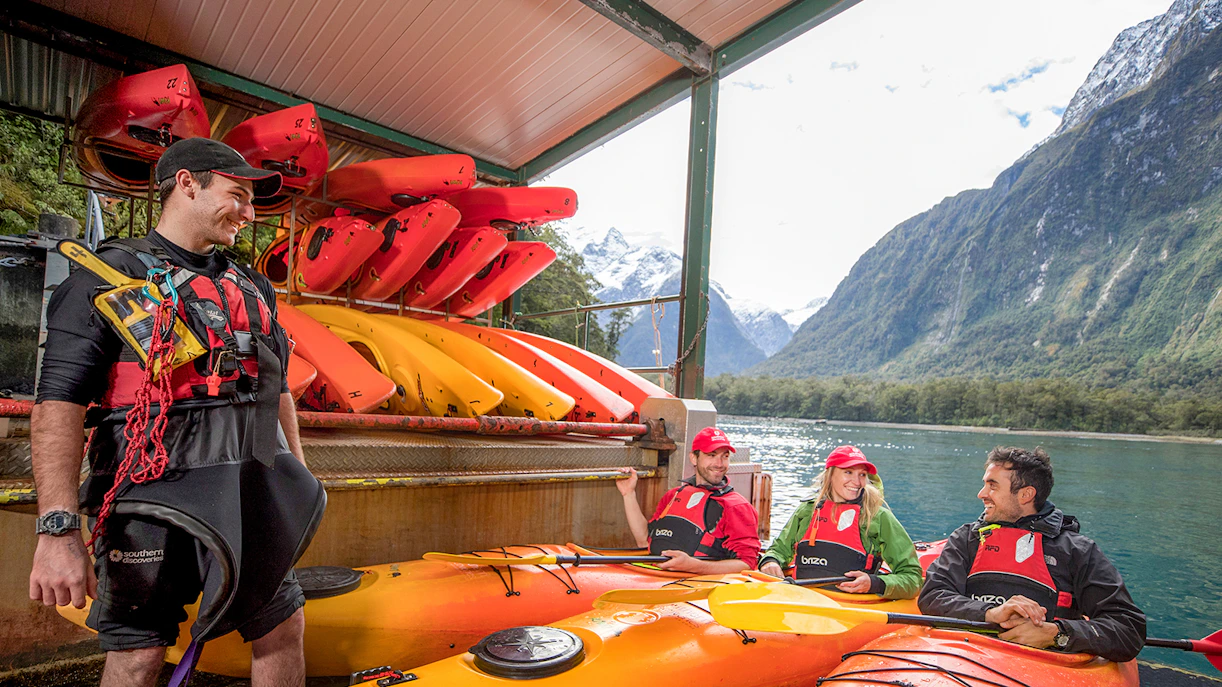 Kayak guide briefing group in Milford Sound with kayaks and mountains in background.