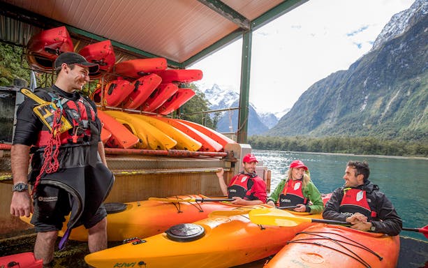 Kayak guide briefing group in Milford Sound with kayaks and mountains in background.