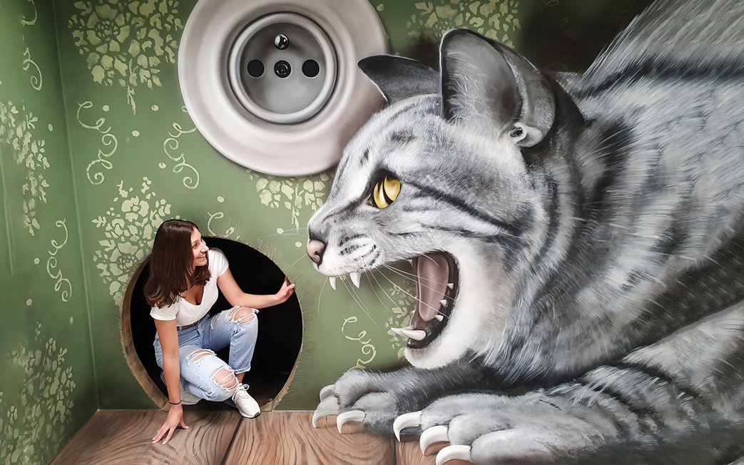 Person crouching in a tunnel next to a giant cat mural at the Museum of Fantastic Illusions.