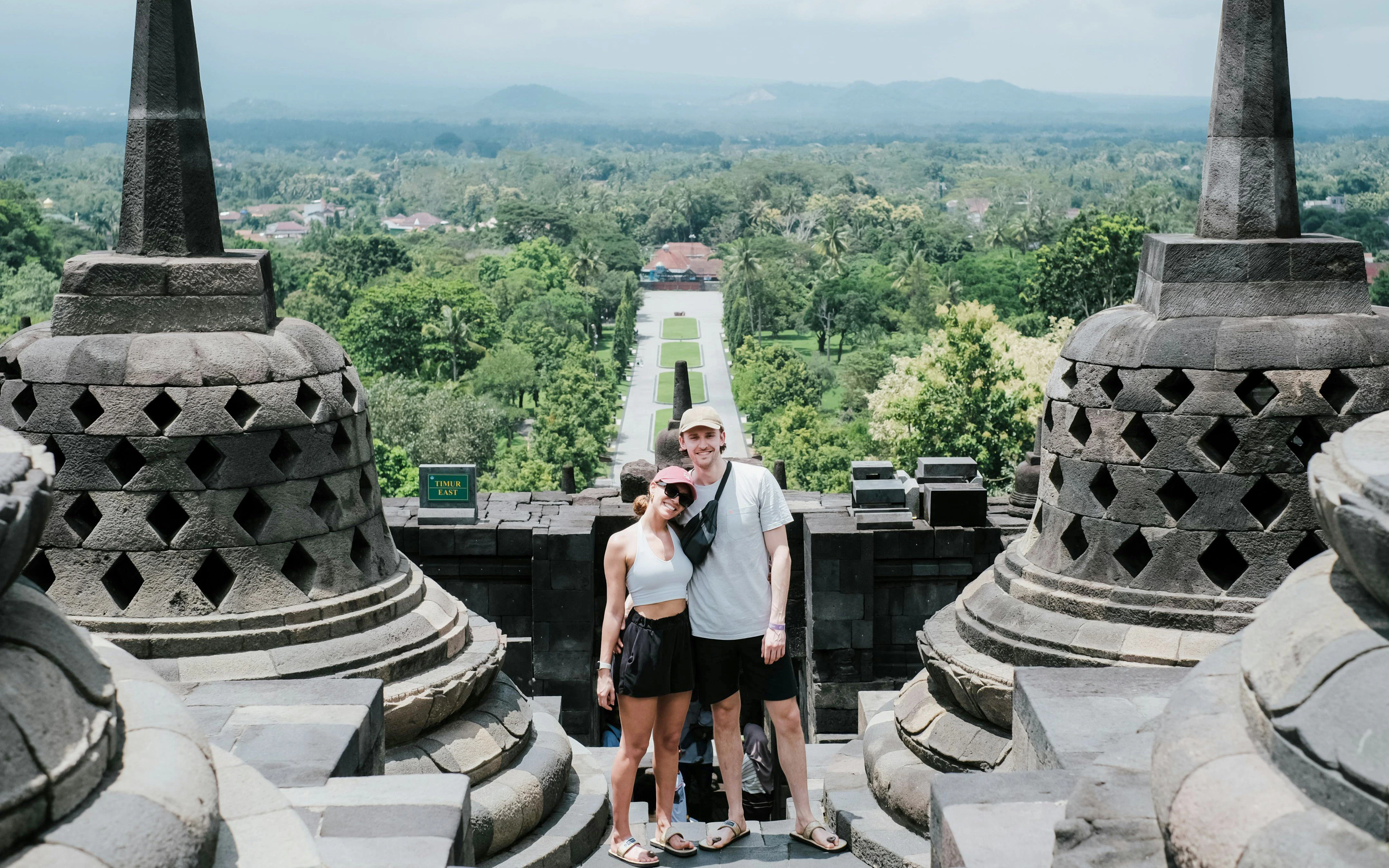 Couple standing at Borobudur Temple with lush landscape in the background.