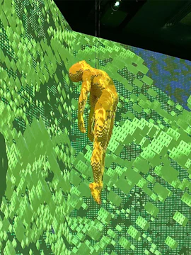 Yellow LEGO figure suspended against a green pixelated background at Art of the Brick exhibit.