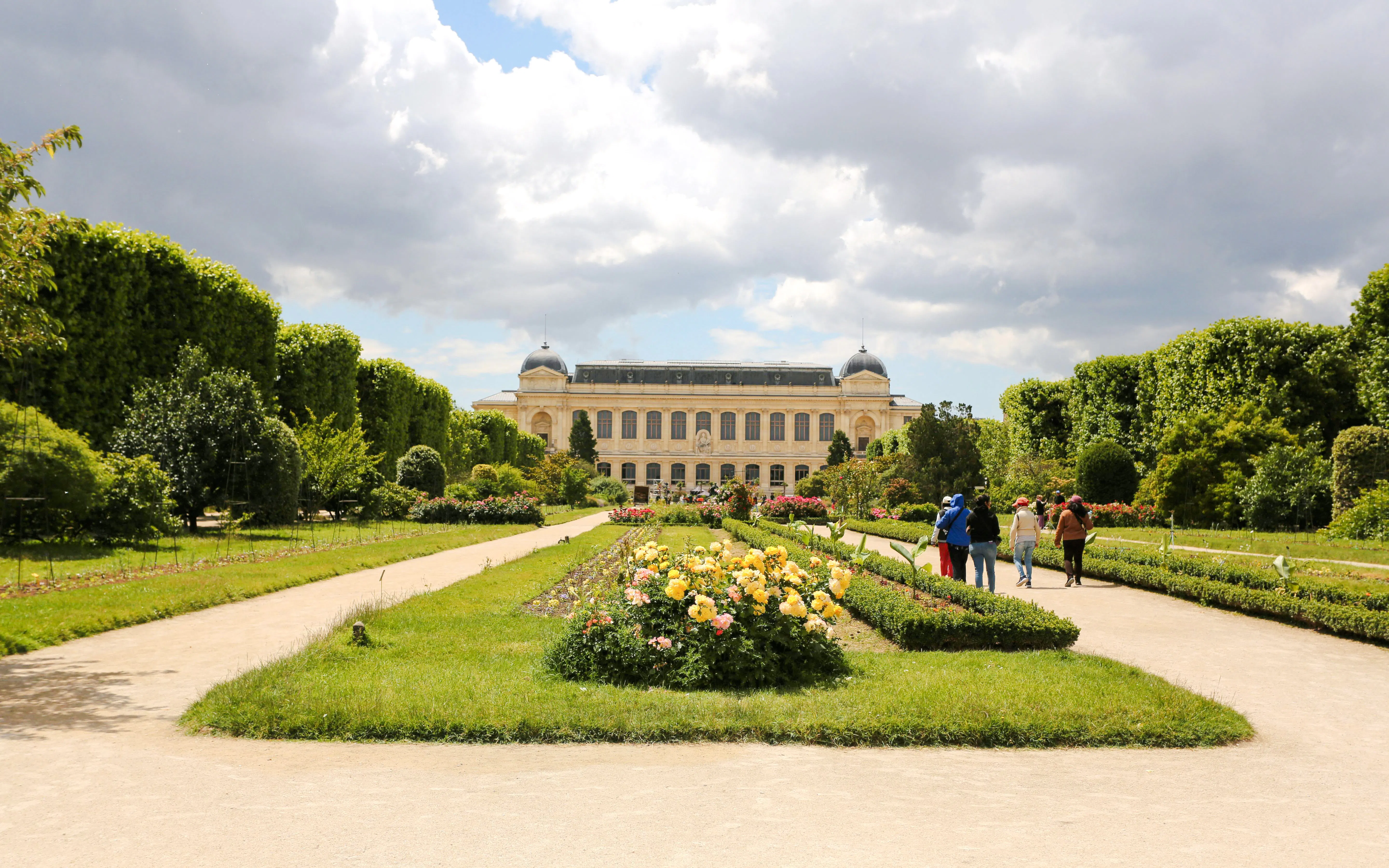 Jardin des Plantes Paris with people walking along flower-lined paths toward a historic building.