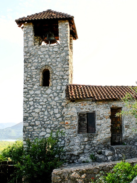 Stone bell tower of the Kom Monastery, Lake Skadar, surrounded by greenery.