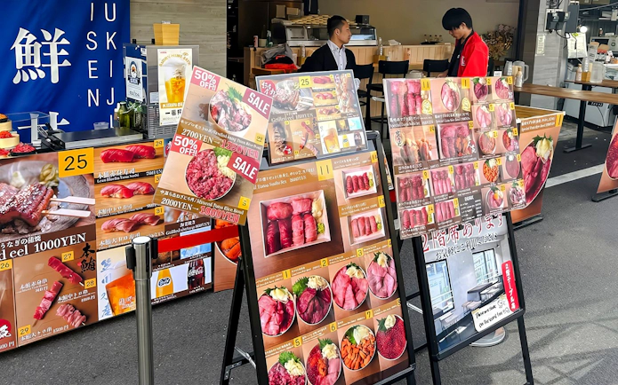 Food stall menu displays at Tsukiji Market, featuring sushi and seafood options.