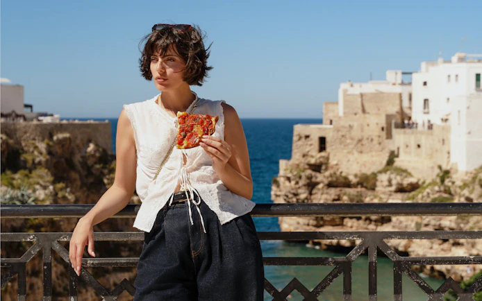 Tourist enjoying pizza with a view of Polignano a Mare coastline.