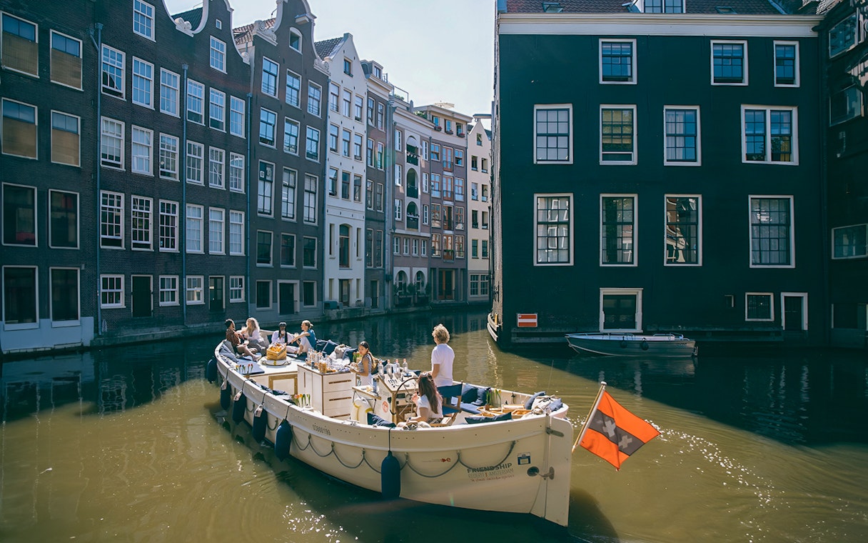 Boat cruising Amsterdam canal with people enjoying drinks, surrounded by historic buildings.