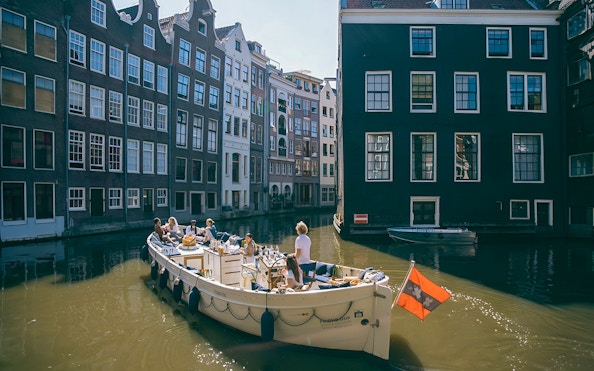 Boat cruising Amsterdam canal with people enjoying drinks, surrounded by historic buildings.