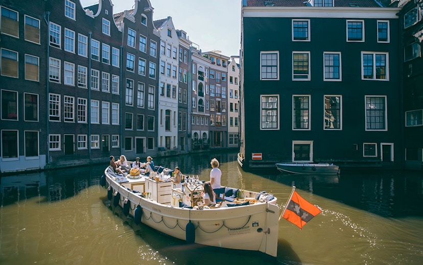 Boat cruising Amsterdam canal with people enjoying drinks, surrounded by historic buildings.