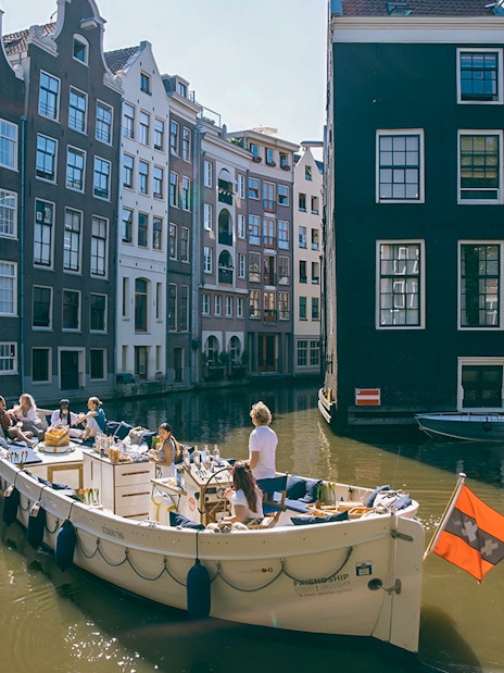 Boat cruising Amsterdam canal with people enjoying drinks, surrounded by historic buildings.