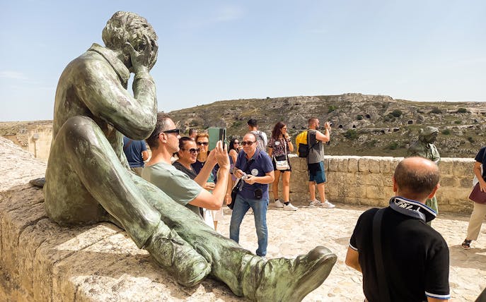 Tourists photographing a statue on a Matera bus tour with views of ancient cave dwellings.