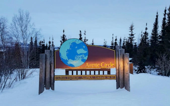 Arctic Circle sign on Dalton Highway, Alaska, surrounded by snow and trees at dusk.