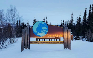 Arctic Circle sign on Dalton Highway, Alaska, surrounded by snow and trees at dusk.