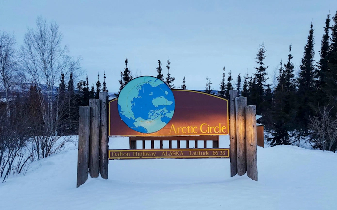 Arctic Circle sign on Dalton Highway, Alaska, surrounded by snow and trees at dusk.