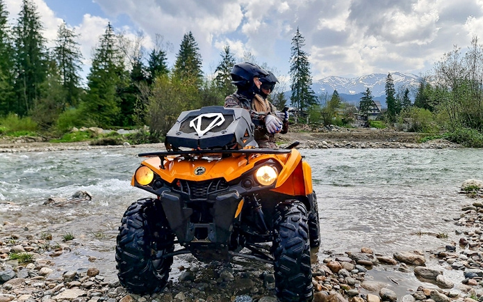Visitors riding a quad bike by a river in Zakopane with mountains in the background.