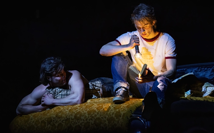 Two young men sitting on a bed, one reading with a flashlight, scene from "The Outsiders.