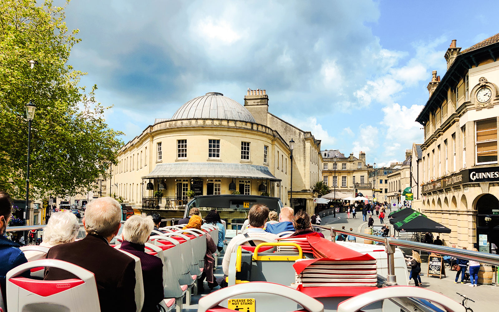 Open-top bus tour near Sheldonian Theatre, Oxford, with passengers viewing historic architecture.