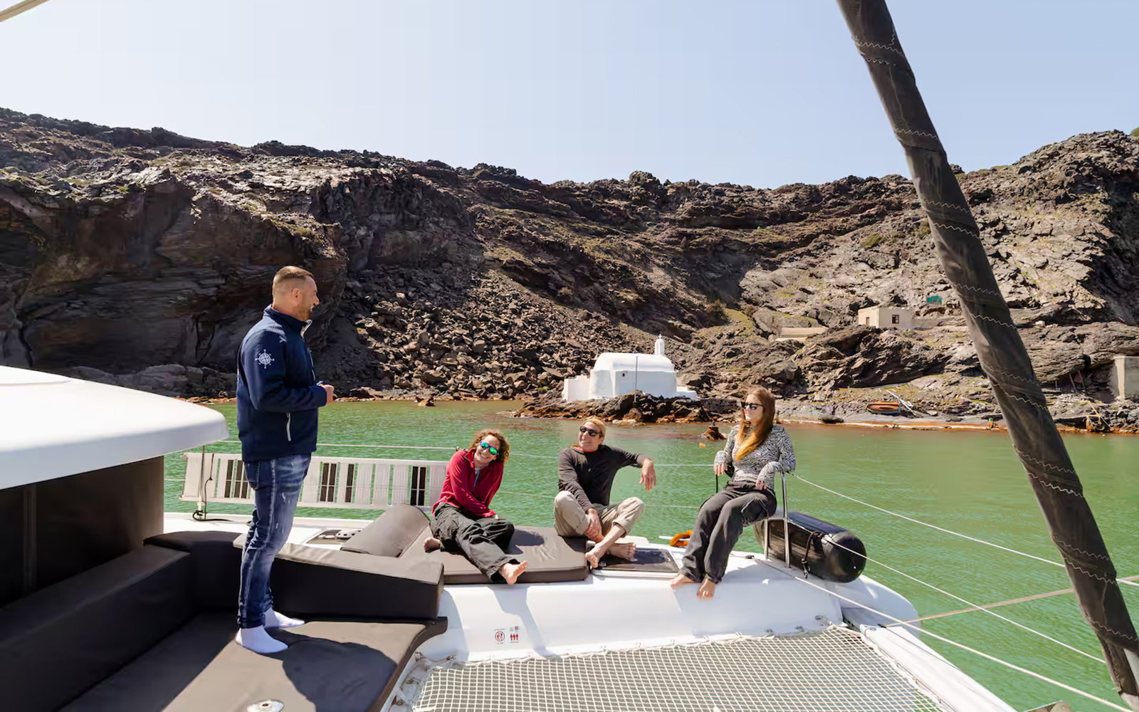 Tour guide with guests on catamaran during Santorini Joy Daytime Cruise, rocky coastline in background.