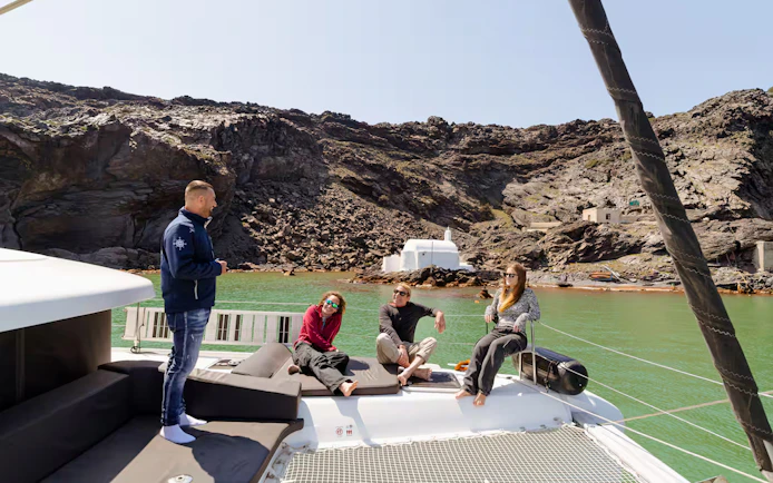 Tour guide with guests on catamaran during Santorini Joy Daytime Cruise, rocky coastline in background.
