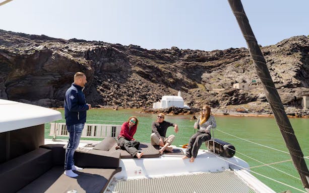 Tour guide with guests on catamaran during Santorini Joy Daytime Cruise, rocky coastline in background.