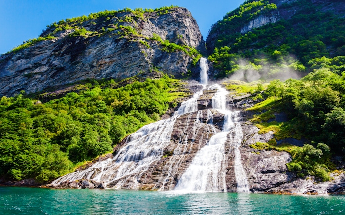 Friaren Waterfall cascading down a rocky cliff surrounded by lush greenery in Geiranger, Norway.
