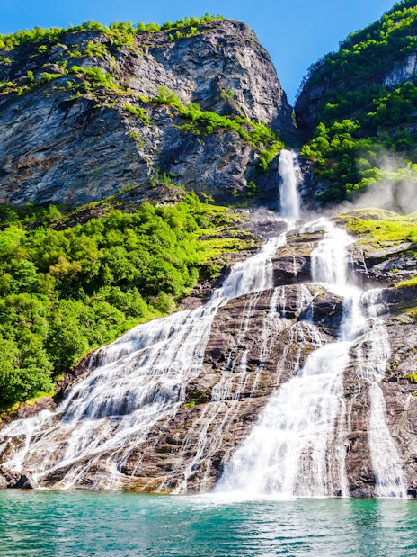 Friaren Waterfall cascading down a rocky cliff surrounded by lush greenery in Geiranger, Norway.