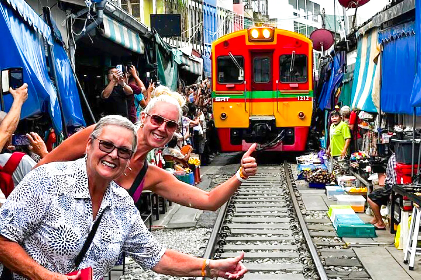 Train passing through Maeklong Railway Market with tourists taking photos, part of the Floating Market & Railway Market tour.
