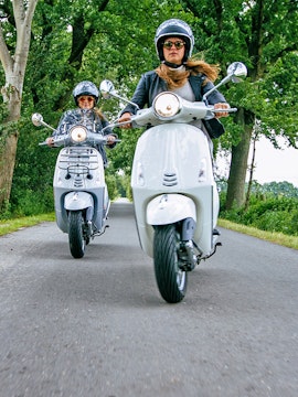 Two people riding scooters on a tree-lined road.