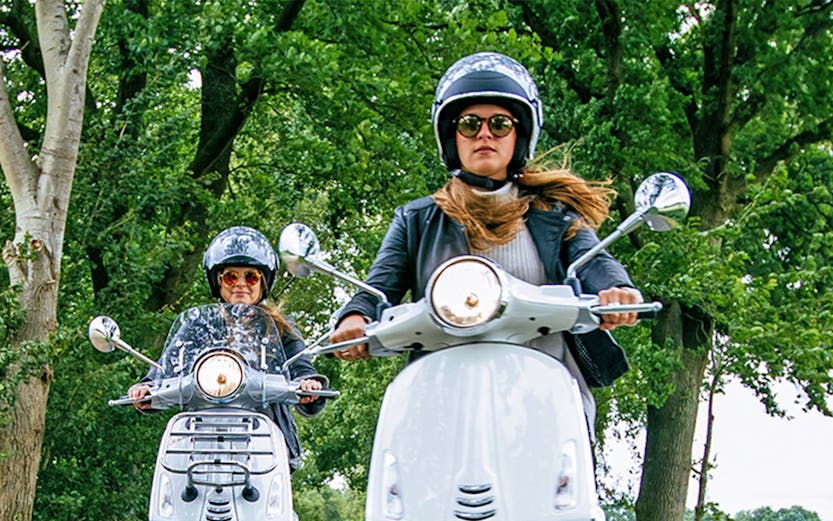 Two people riding scooters on a tree-lined road.