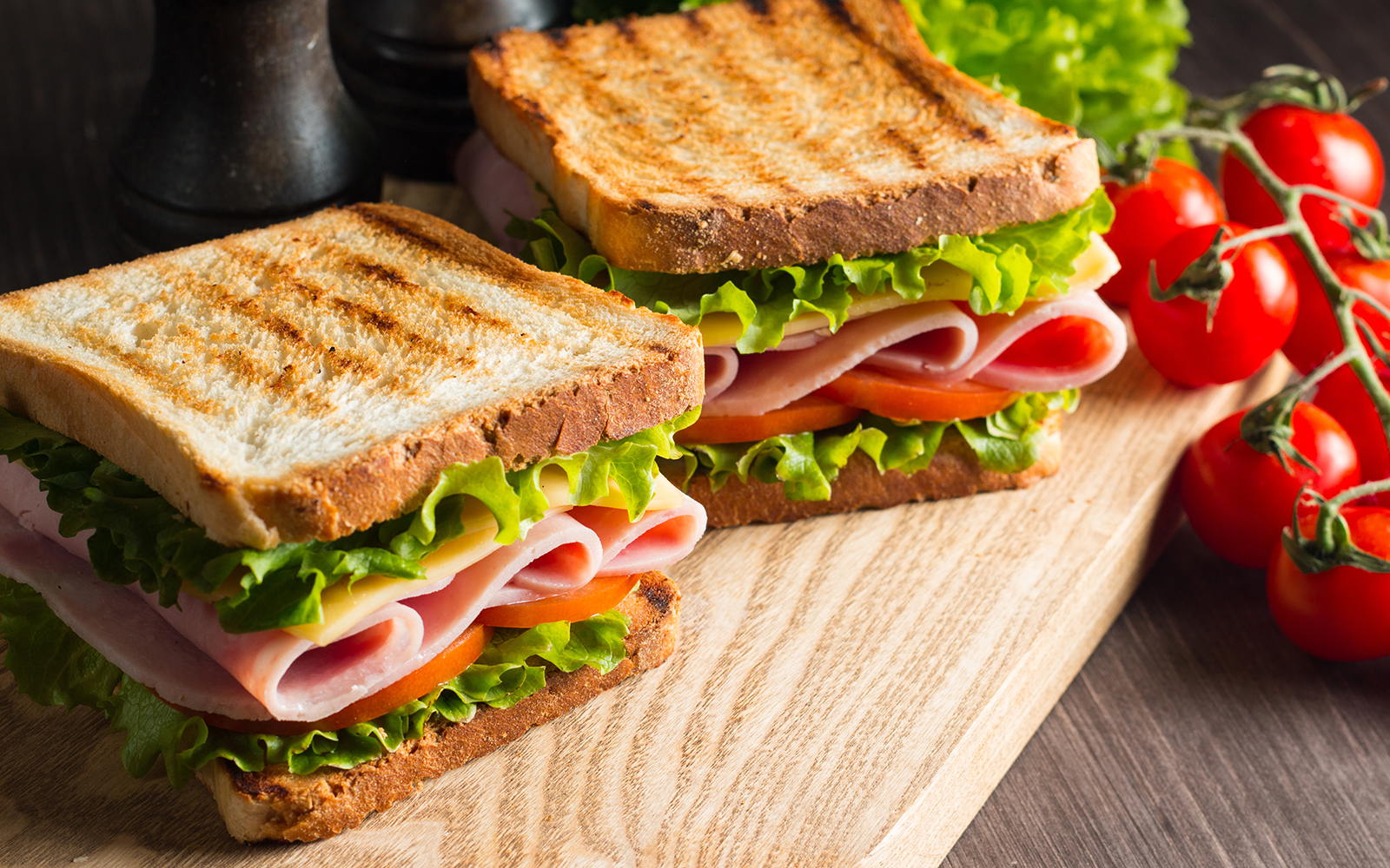 Ham and cheese sandwiches with lettuce and tomato on a wooden board, next to vine tomatoes.