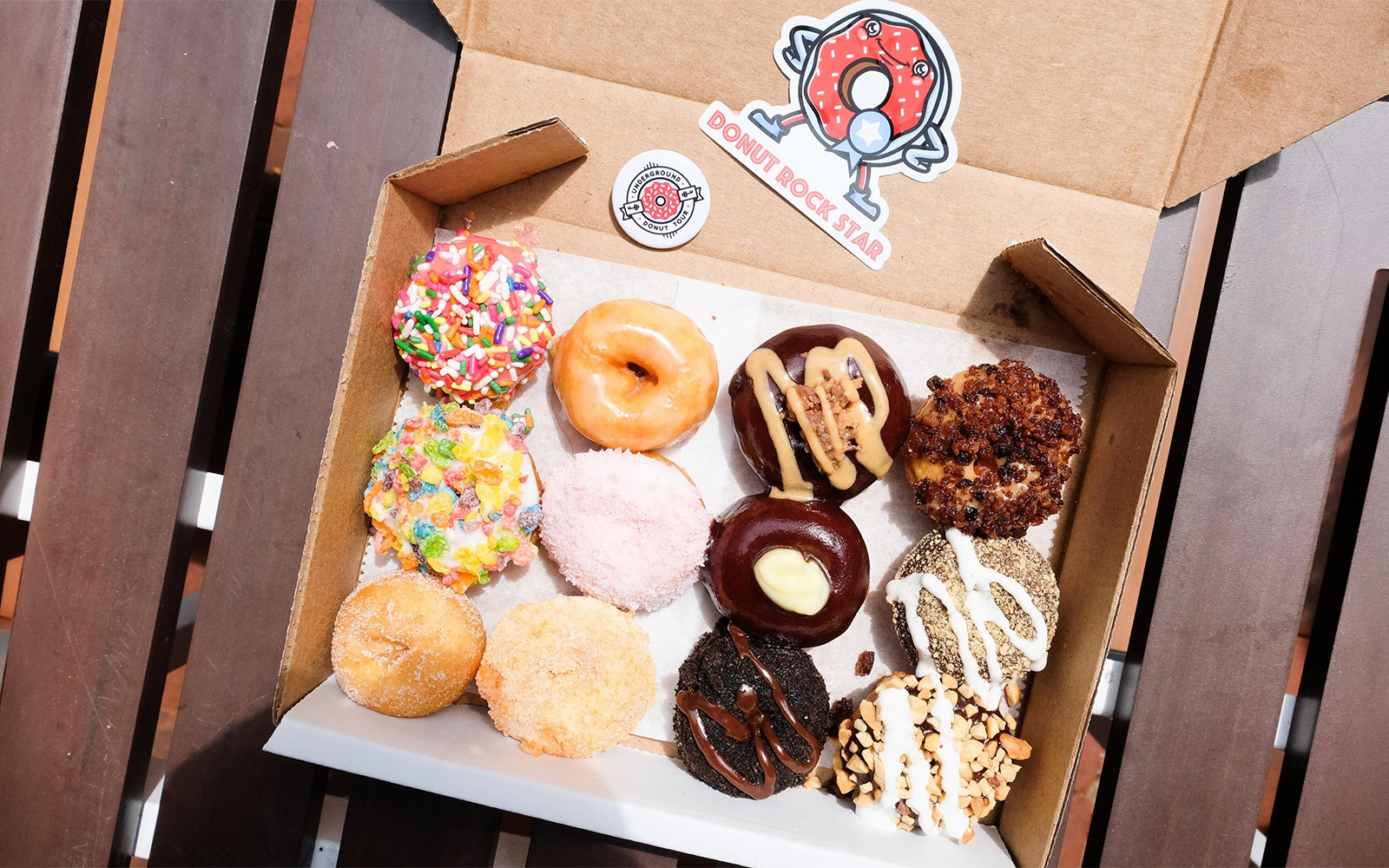 Assorted donuts with various toppings in a box on a wooden table.