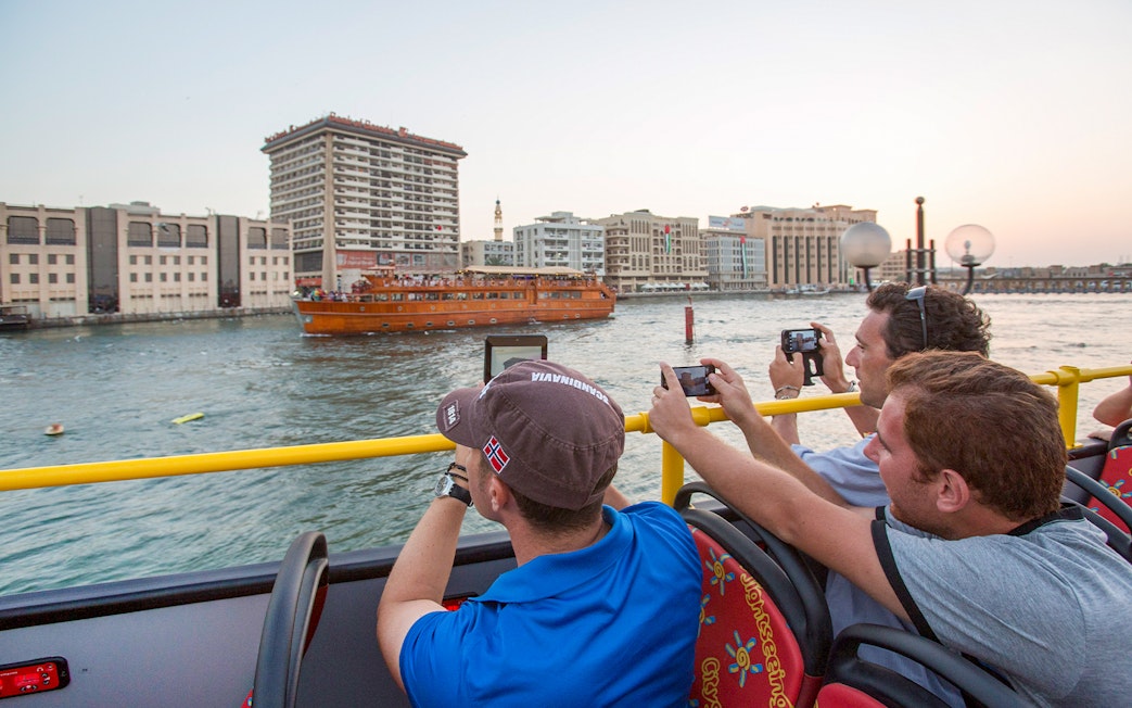Tourists on HOHO bus photographing dhow cruise on Dubai Creek.
