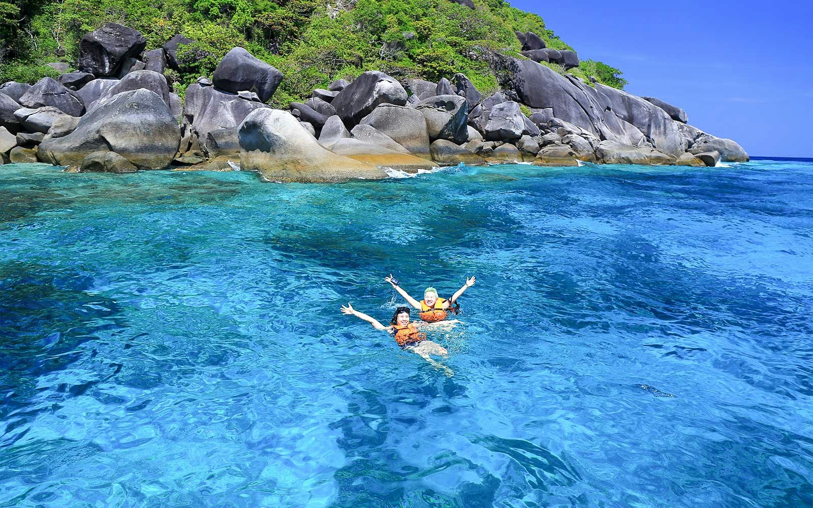 Two people swimming in clear blue water near rocky shore, Similan Islands tour.