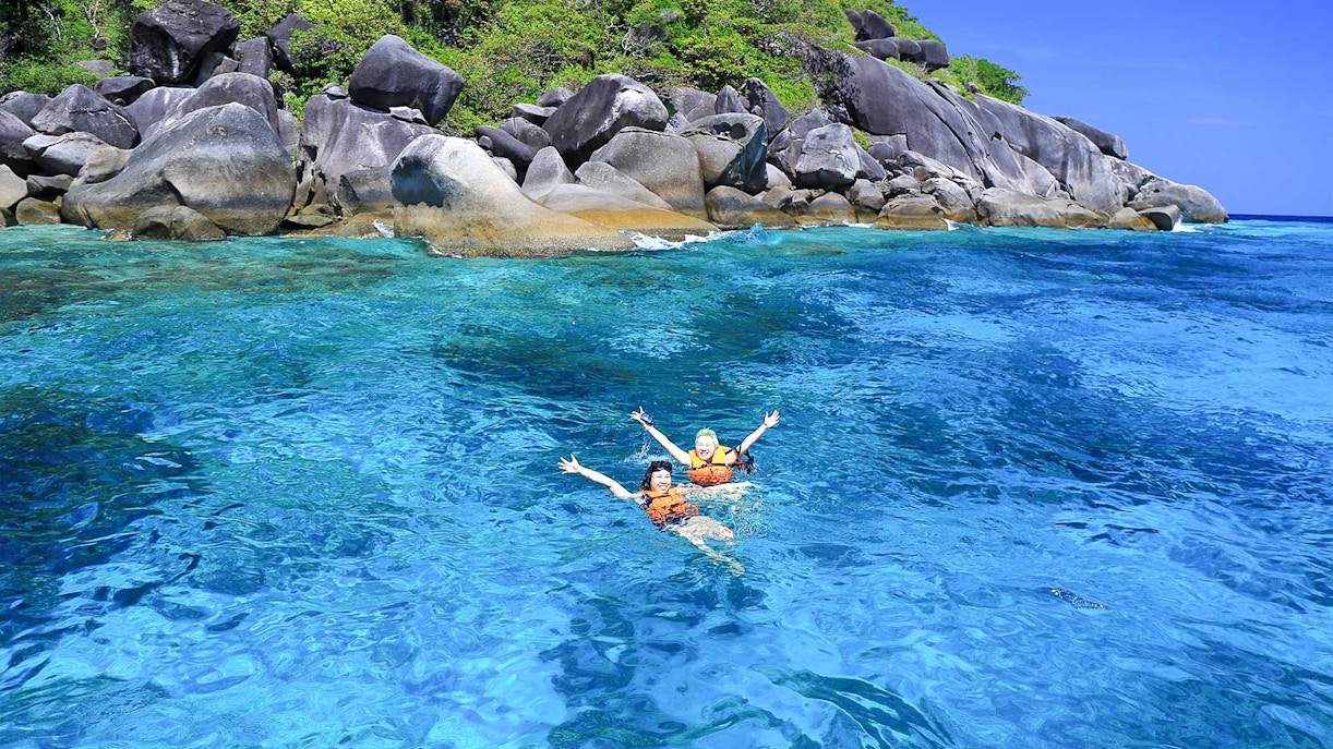 Two people swimming in clear blue water near rocky shore, Similan Islands tour.