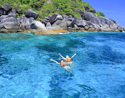 Two people swimming in clear blue water near rocky shore, Similan Islands tour.