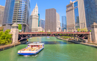 Chicago River cruise boat passing under a bridge with city skyscrapers in the background.