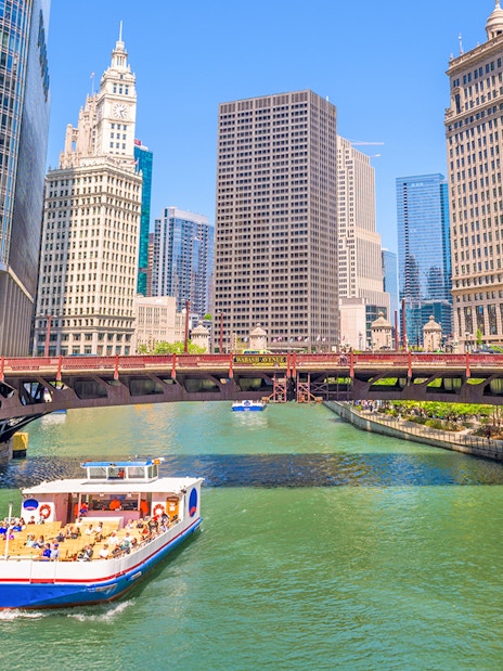 Chicago River cruise boat passing under a bridge with city skyscrapers in the background.