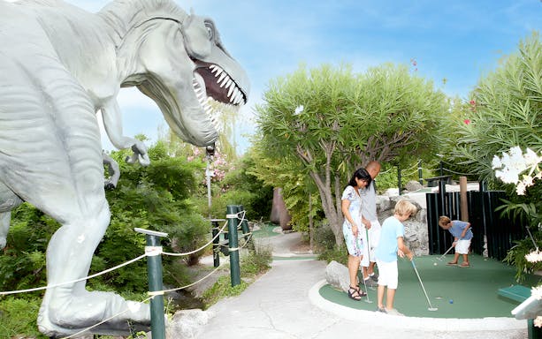 Family playing mini-golf near dinosaur statue at adventure park.