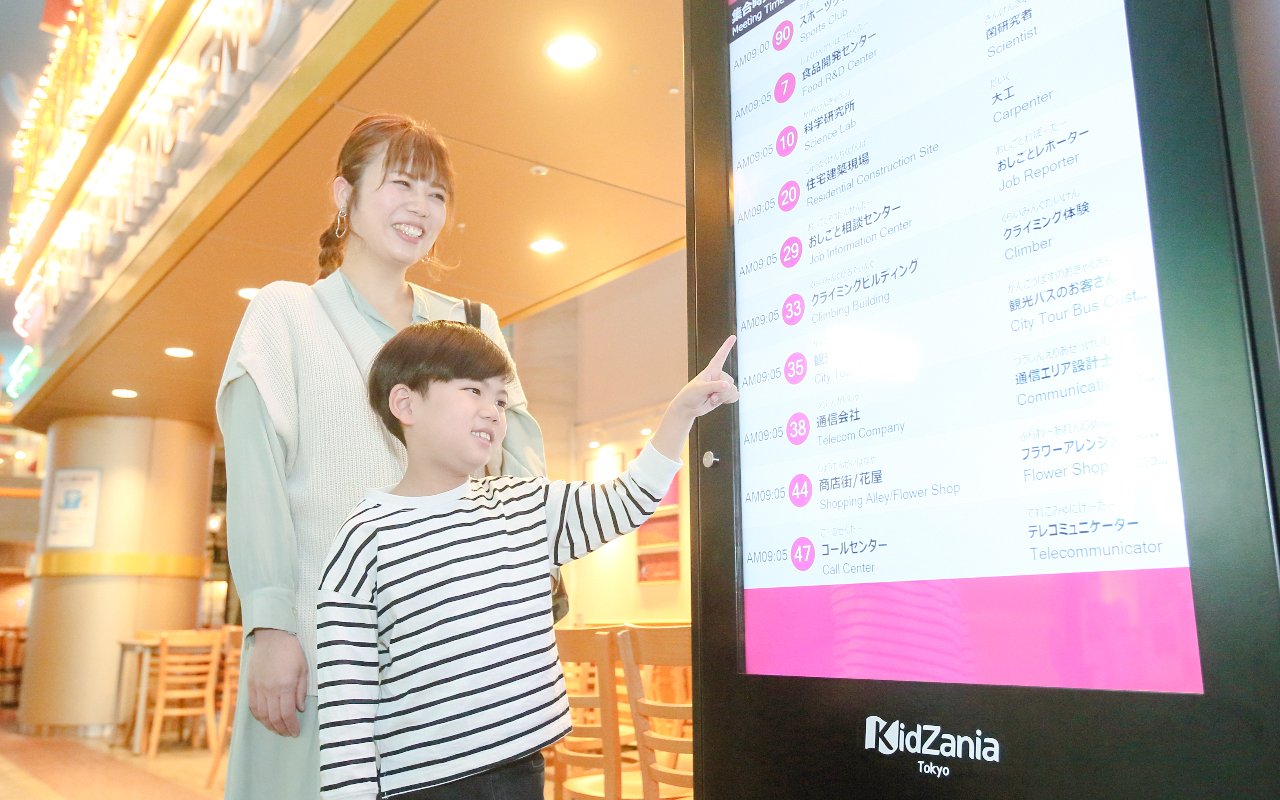 Child pointing at KidZania Tokyo schedule with mother nearby.