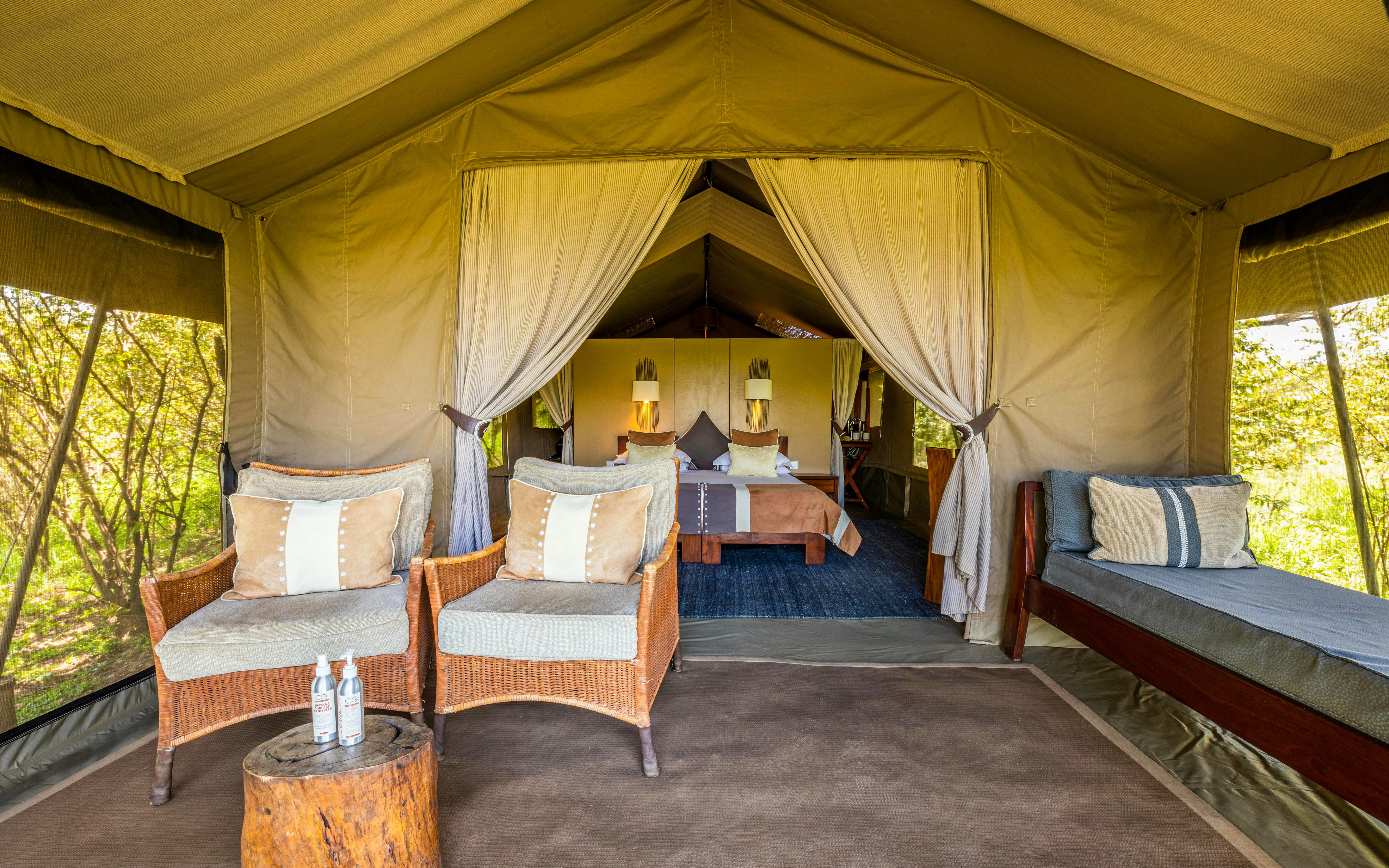 Safari tent interior with seating area and bed, surrounded by lush greenery.