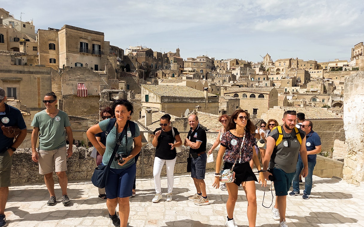 Tourists exploring Matera with audio guide, historic cave houses in background.