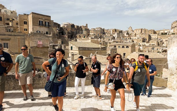Tourists exploring Matera with audio guide, historic cave houses in background.