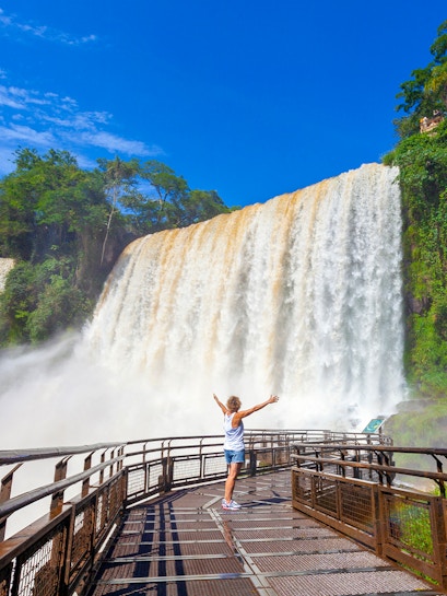 Visitor on walkway admiring Iguazu Falls, surrounded by lush greenery.
