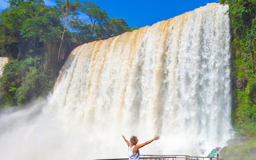 Visitor on walkway admiring Iguazu Falls, surrounded by lush greenery.