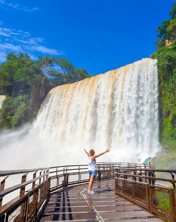 Visitor on walkway admiring Iguazu Falls, surrounded by lush greenery.