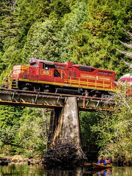 Red California Western train crossing a bridge over a river with kayakers below, surrounded by lush forest in San Francisco.