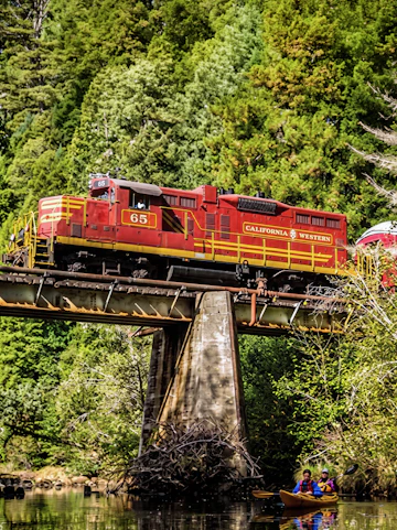 Red California Western train crossing a bridge over a river with kayakers below, surrounded by lush forest in San Francisco.