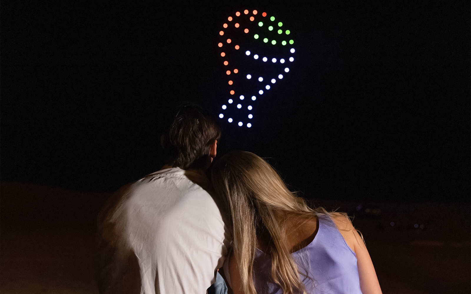 Couple watching drone light show forming hot air balloon shape in Dubai desert.